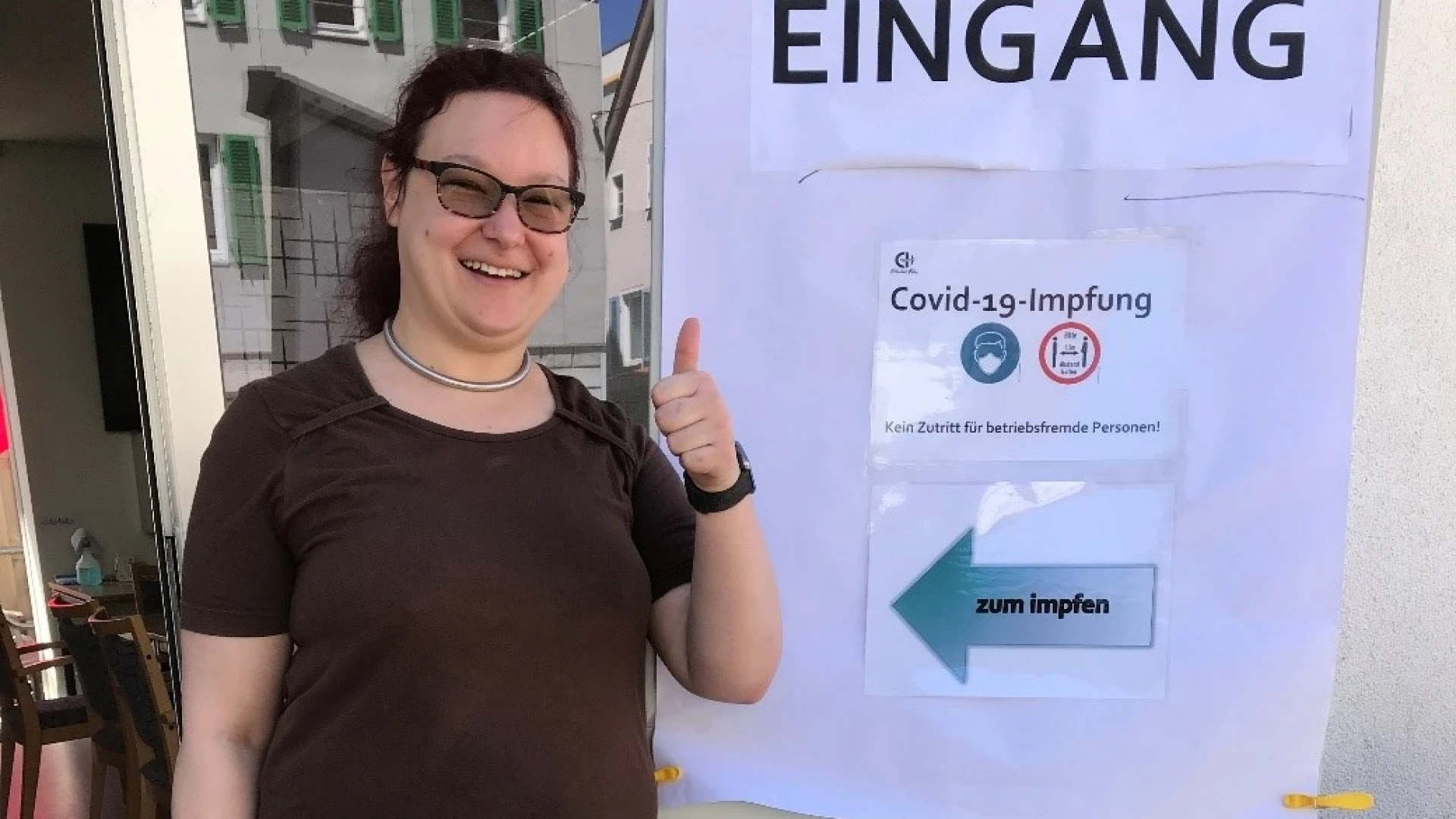 Eine Frau steht vor einem Eingangsschild mit der Aufschrift "Covid-19-Impfung" und h&auml;lt ihren Daumen in die H&ouml;he. Ein Pfeil auf dem Plakat weist auf den Impfbereich hin.