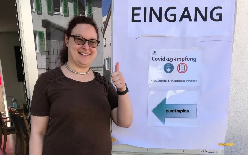 Eine Frau steht vor einem Eingangsschild mit der Aufschrift "Covid-19-Impfung" und h&auml;lt ihren Daumen in die H&ouml;he. Ein Pfeil auf dem Plakat weist auf den Impfbereich hin.