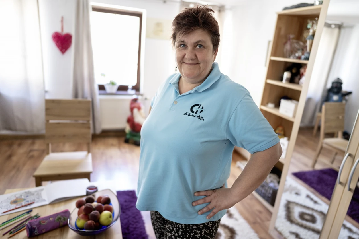 Eine Frau in hellblauem Polo-Shirt mit EH Logo steht l&auml;chelnd in einem Raum mit Holzboden, B&uuml;cherregal und Obstschale. Sie hat ihre H&auml;nde in die H&uuml;ften gest&uuml;tzt.