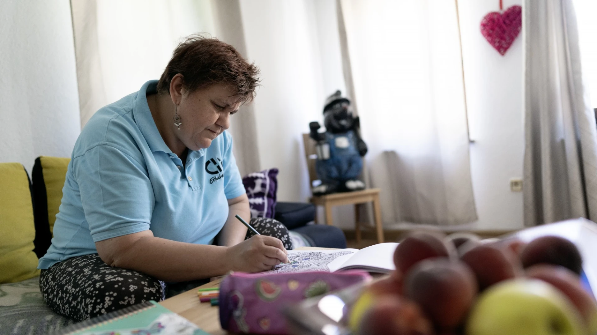 Eine Frau in blauem Poloshirt und EH Logo sitzt an einem Tisch mit B&uuml;chern und Obst, konzentriert mit einem Stift in der Hand malt sie etwas. Im Hintergrund sind Vorh&auml;nge und ein herzf&ouml;rmiges Dekorationselement zu sehen.
