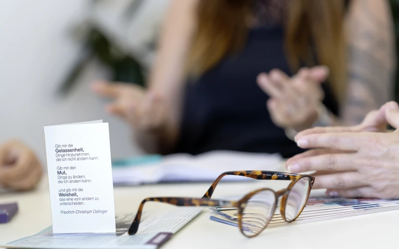 Braune Lesebrille liegt auf einem Tisch neben einem wei&szlig;en Dokument, w&auml;hrend im Hintergrund verschwommen eine Person sitzt und Handgesten macht.