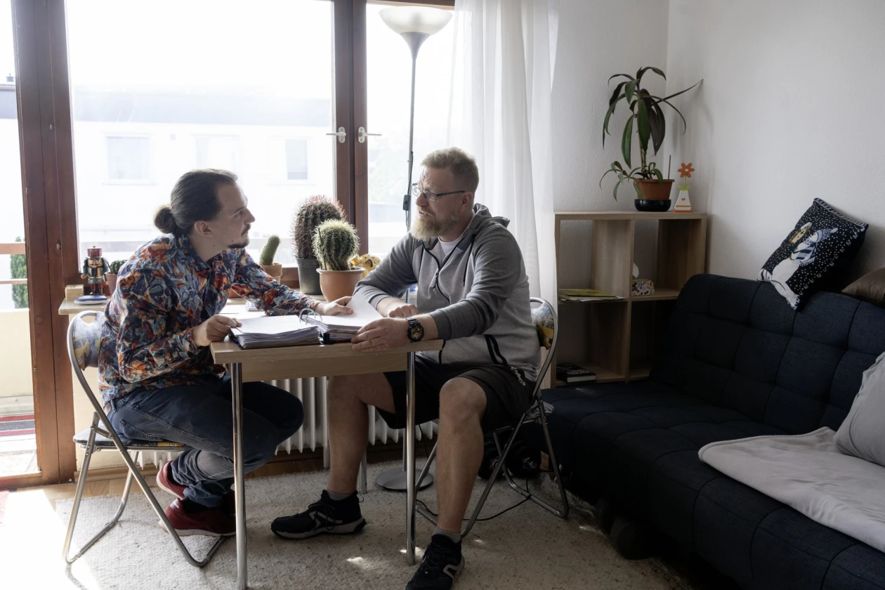 Ein Mitarbeiter ist mit einem Mann im Gespr&auml;ch an einem Tisch mit Ordner, in einem hellen Raum mit Panoramafenster. Der Raum ist mit einem dunkelblauen Sofa und einem Regal eingerichtet.