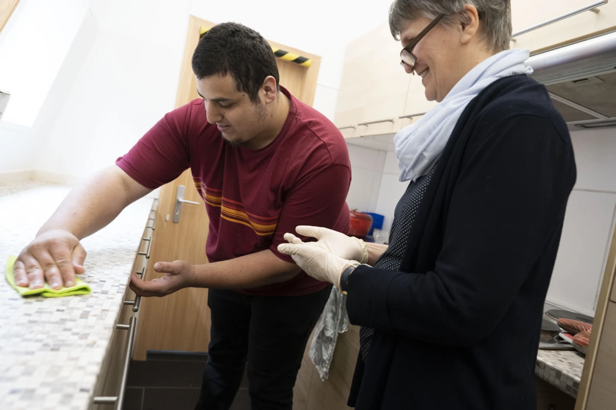 Ein Mann in einem roten T-Shirt und eine Mitarbeiterin stehen gemeinsam in einer K&uuml;che. Der Mann wischt eine Arbeitsfl&auml;che ab.