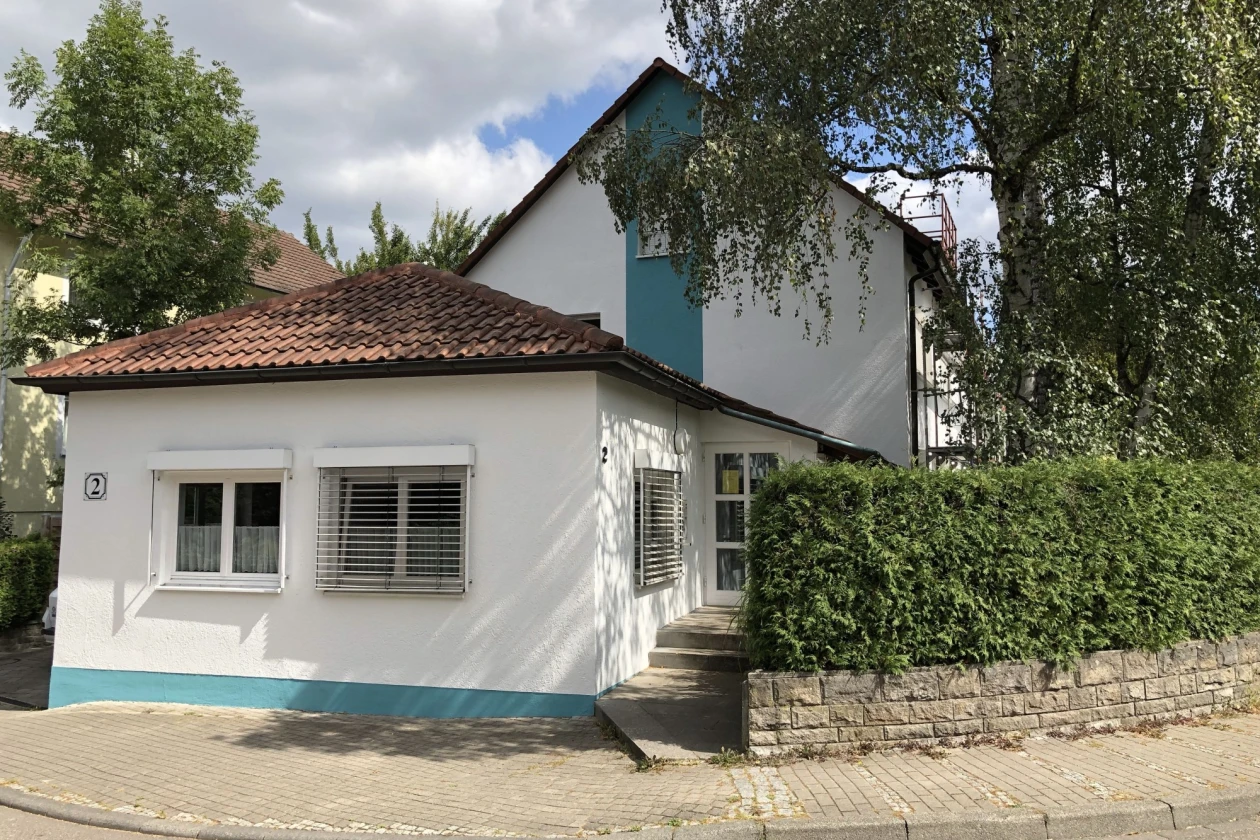 Wei&szlig;es Einfamilienhaus mit t&uuml;rkisfarbenen Akzenten an Sockel und Fassade, umgeben von einer Hecke und B&auml;umen, an einer Stra&szlig;enecke mit Gehweg und Bordstein gelegen.