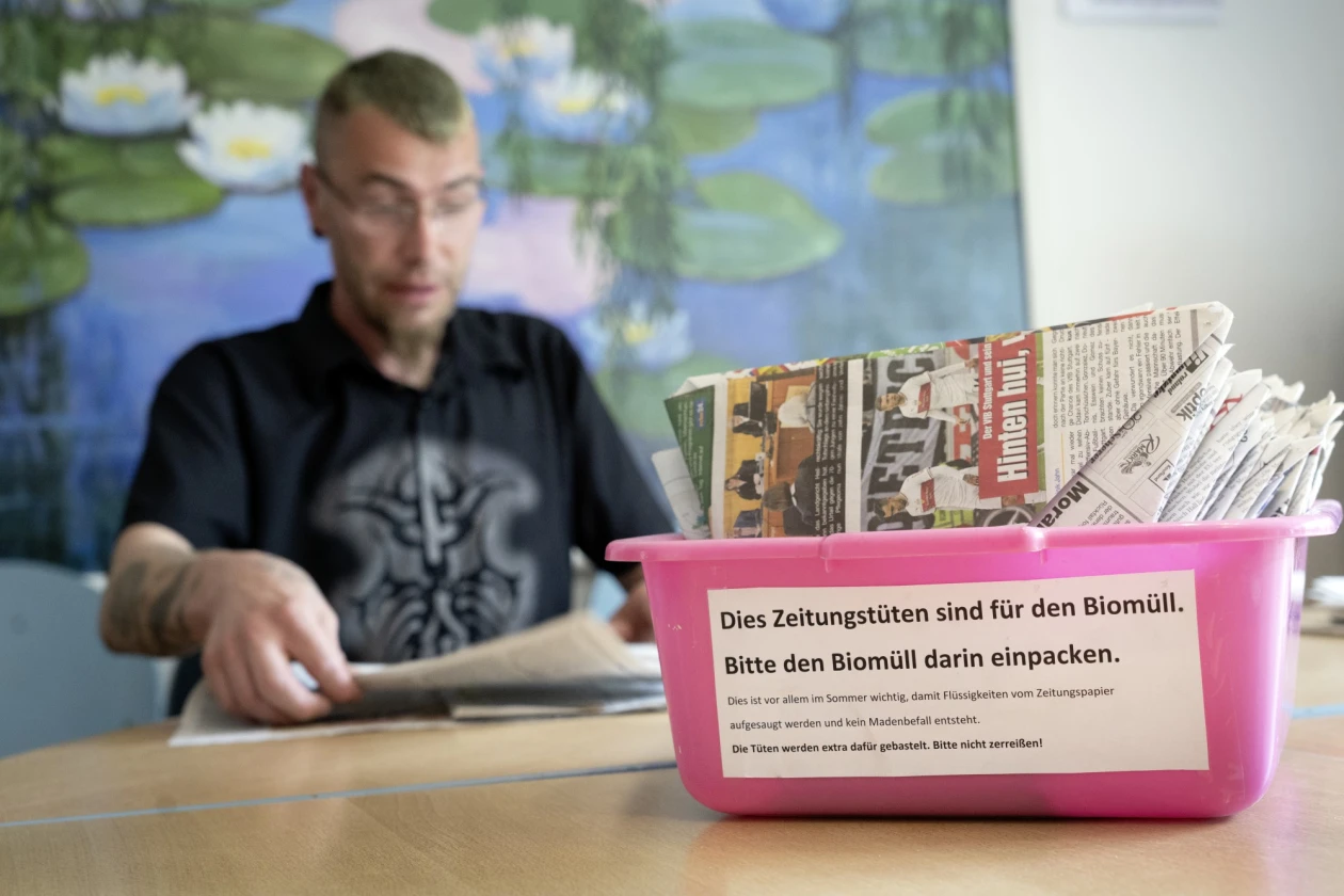 Ein Mann mit schwarzem T-Shirt sitzt an einem Tisch und faltet Zeitungst&uuml;ten, die er in einer pinkfarbenen Box sammelt. Im Hintergrund ist unscharf ein Wandbild mit Seerosen zu sehen.
