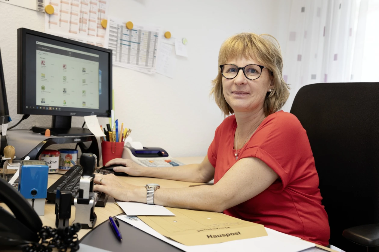 Eine l&auml;chelnde Frau mit Brille im roten T-Shirt sitzt an einem Schreibtisch mit Computer und B&uuml;routensilien.