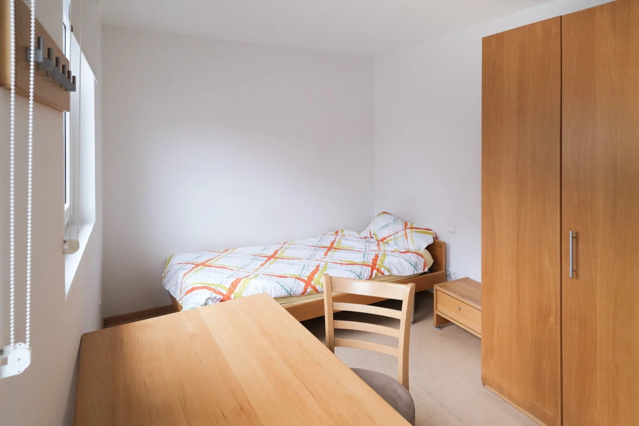 Einzelzimmer mit Holzm&ouml;beln, bestehend aus einem Bett, einem Holzschrank und einem Holztisch mit Stuhl. Das Zimmer hat wei&szlig;e W&auml;nde und eine Jalousie am Fenster, die Bettw&auml;sche ist farbig bezogen.