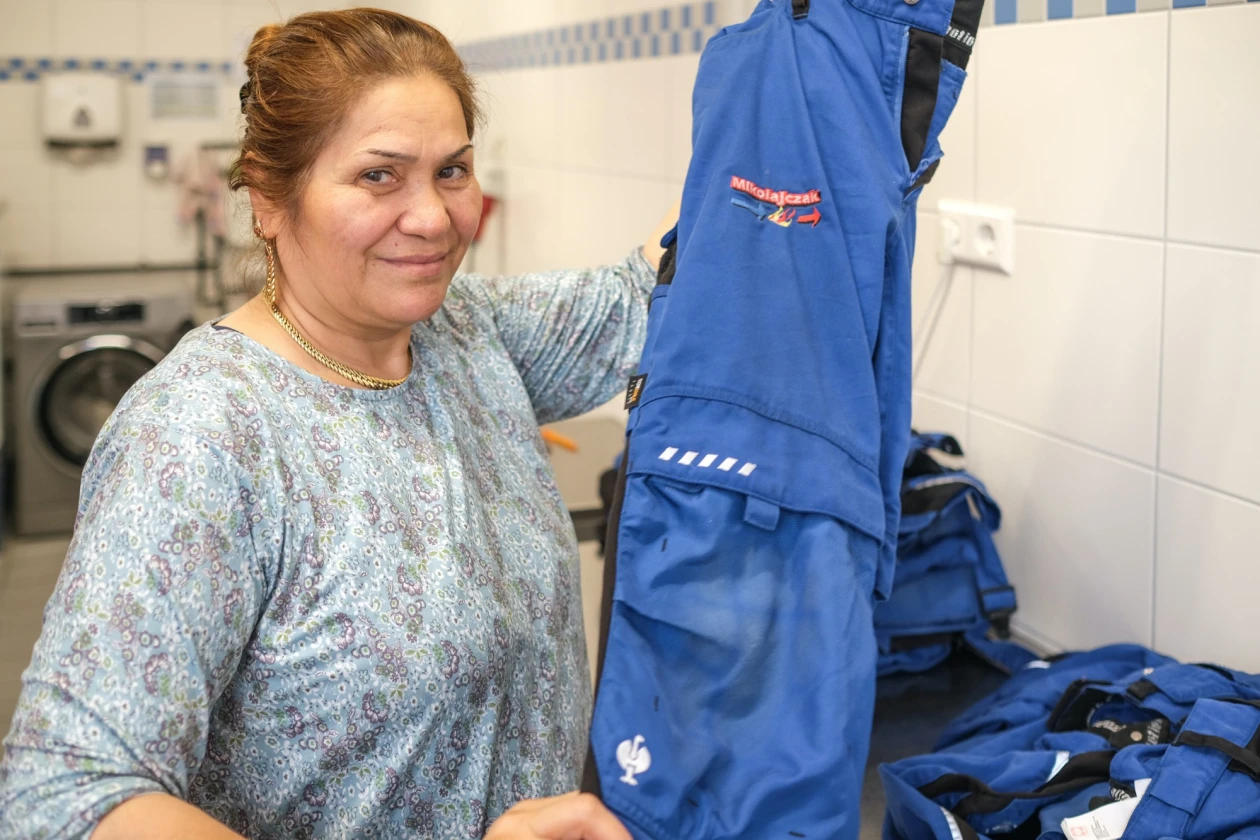 L&auml;chelnde Frau in gemusterter Kleidung steht in einem Waschraum und hebt ein blaues Kleidungsst&uuml;ck in die Kamera.