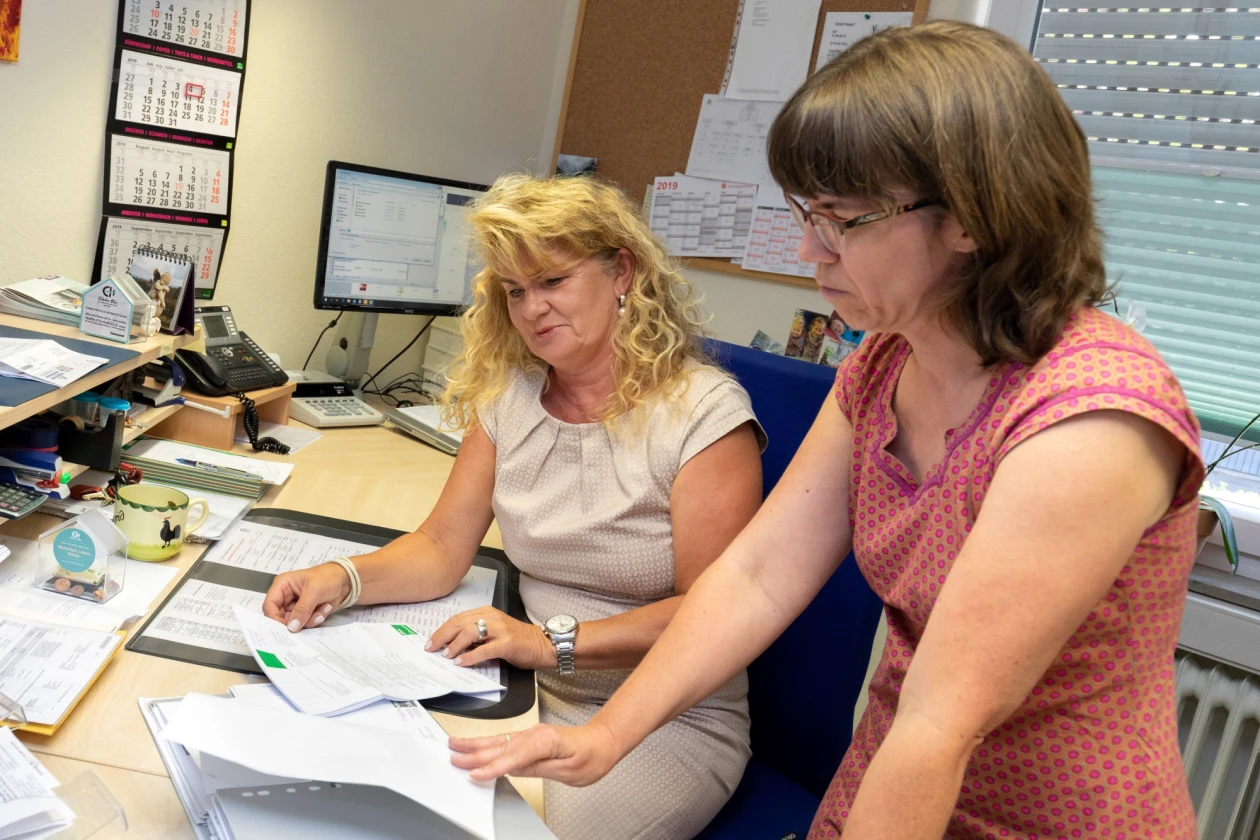 Zwei Frauen arbeiten an einem Schreibtisch in einem B&uuml;ro. Die blondhaarige Frau tr&auml;gt ein beiges Kleid, die braunhaarige Frau tr&auml;gt Brille und ein rosa gepunktetes Kleid.