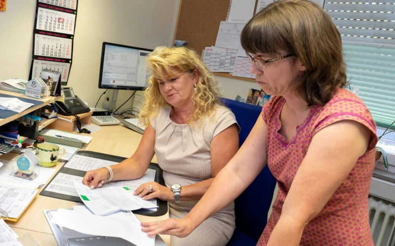Zwei Frauen arbeiten an einem Schreibtisch in einem B&uuml;ro. Die blondhaarige Frau tr&auml;gt ein beiges Kleid, die braunhaarige Frau tr&auml;gt Brille und ein rosa gepunktetes Kleid.