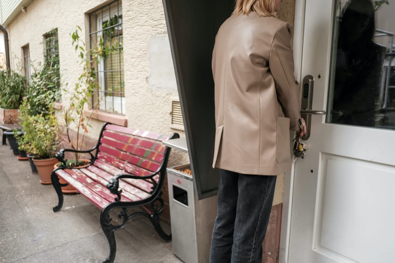 Rote Holzbank an einer Hauswand, daneben Topfpflanzen. Eine Frau in einem beigen Mantel &ouml;ffnet die T&uuml;r mit ihrem Schl&uuml;ssel.