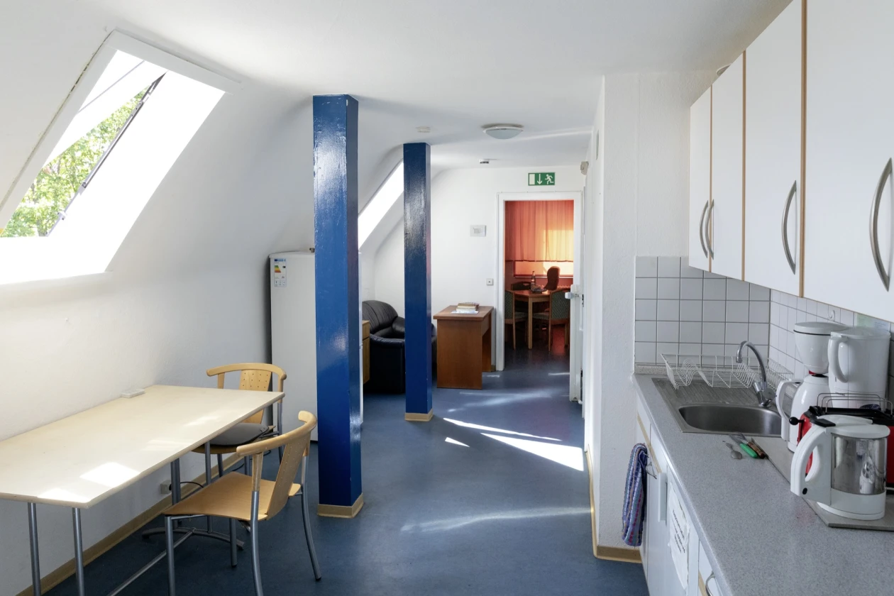 Heller, moderner Gemeinschaftsraum mit K&uuml;che und Essbereich. Wei&szlig;e W&auml;nde, blaue S&auml;ulen und blauer Bodenbelag, Holztisch mit zwei St&uuml;hlen, Kaffeemaschine und Fenster mit Blick ins Gr&uuml;ne. Durchgang zu einem weiteren Raum mit gr&uuml;nem Notausgang-Schild.