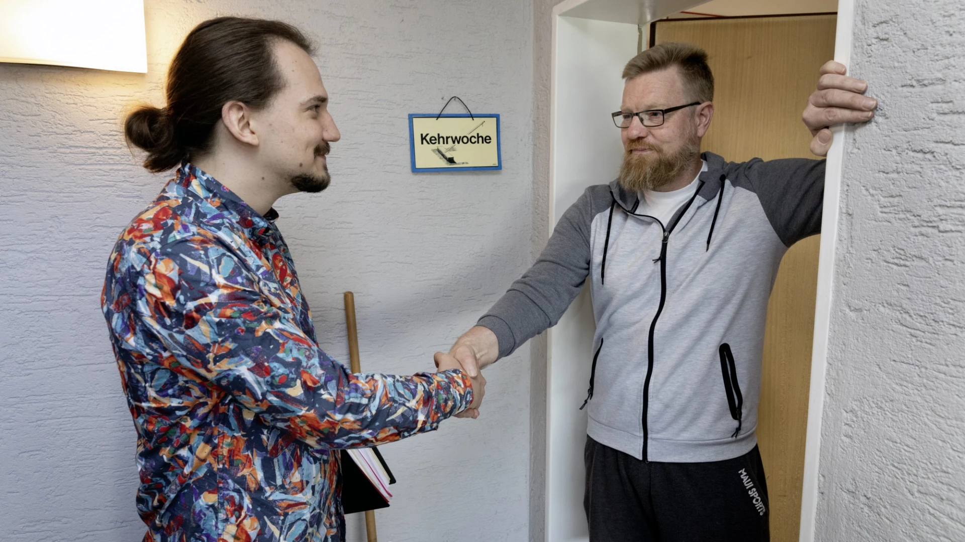 Ein Mitarbeiter und ein Mann sch&uuml;tteln sich in einem Flur mit wei&szlig;en W&auml;nden die H&auml;nde. Der Mitarbeiter tr&auml;gt ein buntes gemustertes Hemd, der andere eine graue Jacke, Brille und schwarze Hose. An der Wand h&auml;ngt ein Schild mit der Aufschrift "Kehrwoche".