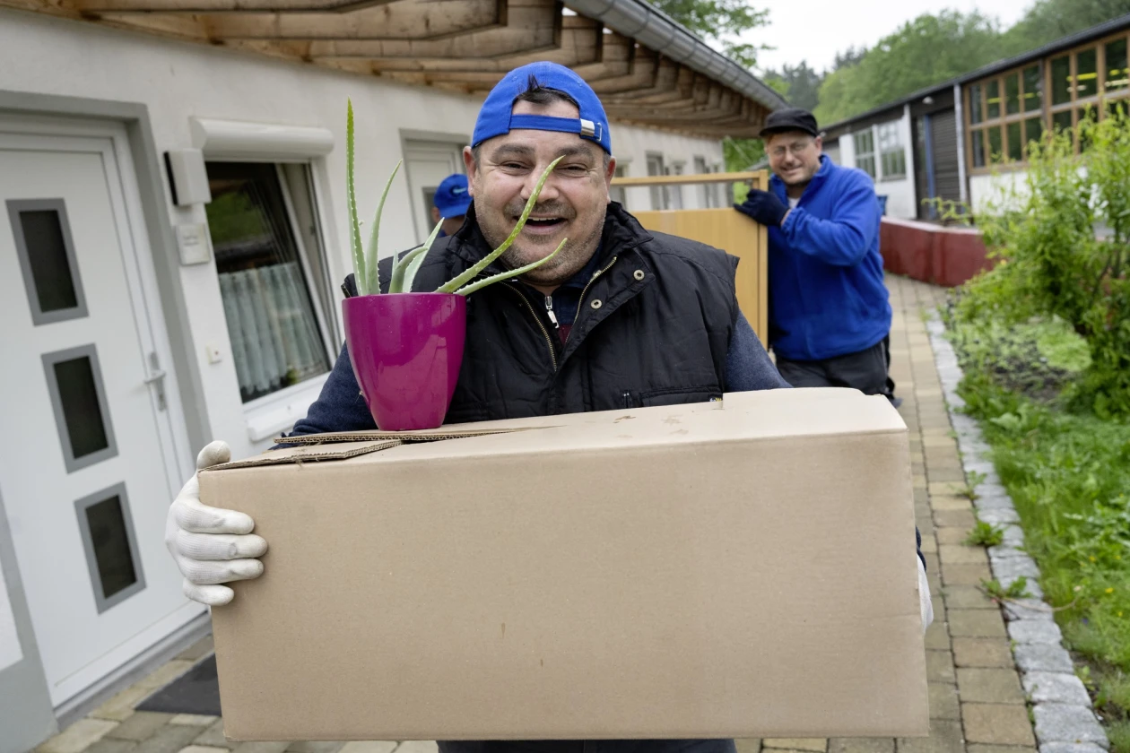 Zwei M&auml;nner tragen Gegenst&auml;nde bei einem Umzug. Der vordere Mann tr&auml;gt eine blaue Kappe und eine schwarze Weste, der hintere Mann eine schwarze Kappe, Brille und eine blaue Jacke.