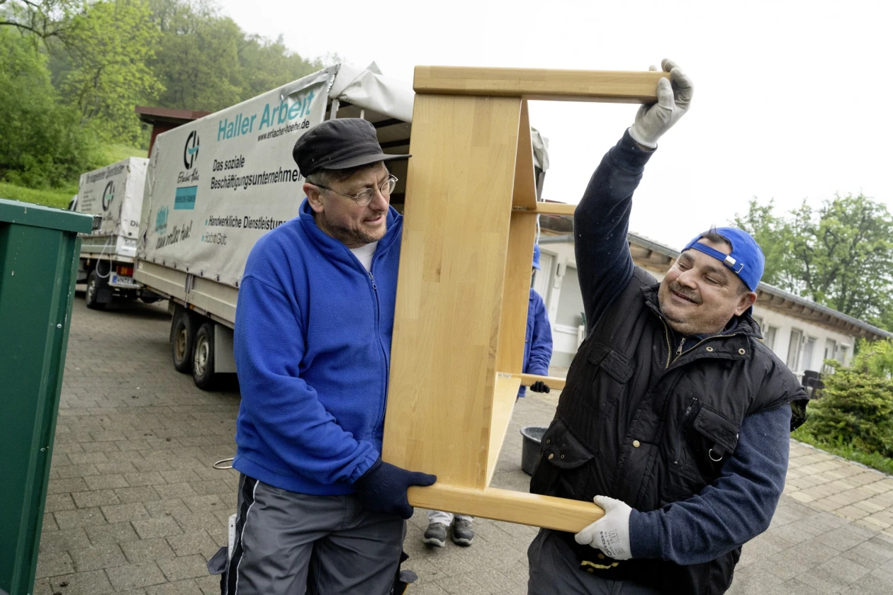 Zwei M&auml;nner in blauer und ein Mann in schwarzer Arbeitskleidung tragen gemeinsam ein Holzbett auf einem Parkplatz, im Hintergrund ist ein Transporter mit Anh&auml;nger der Erlacher H&ouml;he zu sehen.