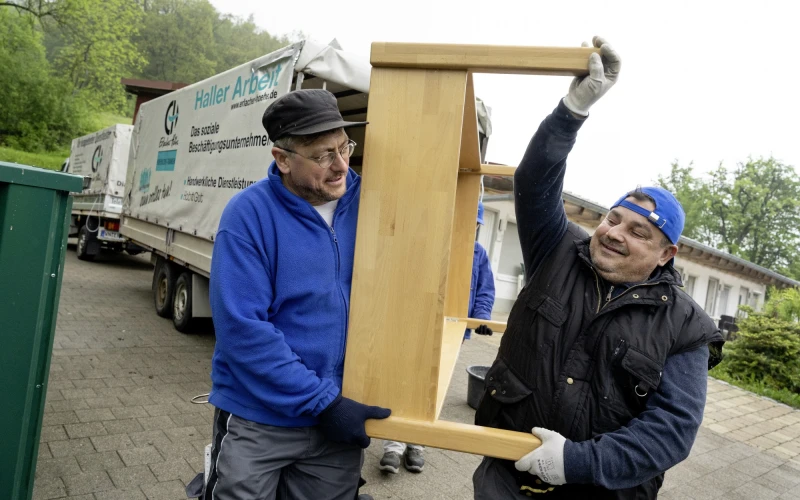 Zwei M&auml;nner in blauer und ein Mann in schwarzer Arbeitskleidung tragen gemeinsam ein Holzbett auf einem Parkplatz, im Hintergrund ist ein Transporter mit Anh&auml;nger der Erlacher H&ouml;he zu sehen.