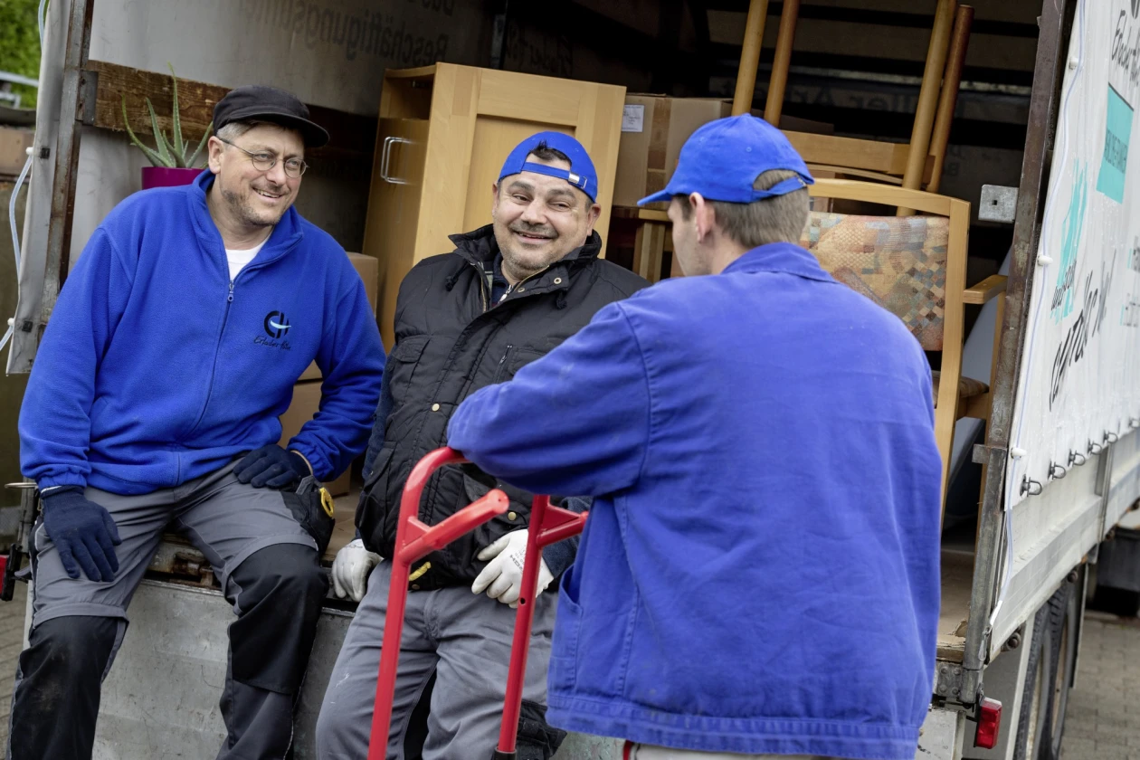 Zu sehen sind zwei M&auml;nner in blauen Jacken und ein Mann mit schwarzer Jacke. Zwei sitzen l&auml;chelnd auf der Ladefl&auml;che eines Transporters, der dritte steht mit dem R&uuml;cken zur Kamera. Im Hintergrund auf der Ladefl&auml;che stehen Schr&auml;nke und St&uuml;hle.