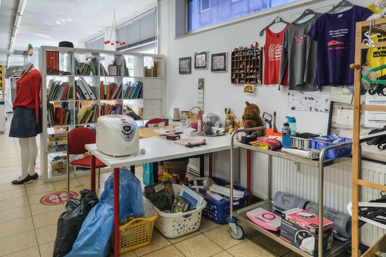 Ein Raum mit B&uuml;cherregalen, Arbeitstisch, Stofftieren, Rollwagen und verschiedenen Alltagsgegenst&auml;nden und Kleidungsst&uuml;cken. Eine Schaufensterpuppe mit einem roten Oberteil und einem grauen Rock steht neben einem B&uuml;cherregal.