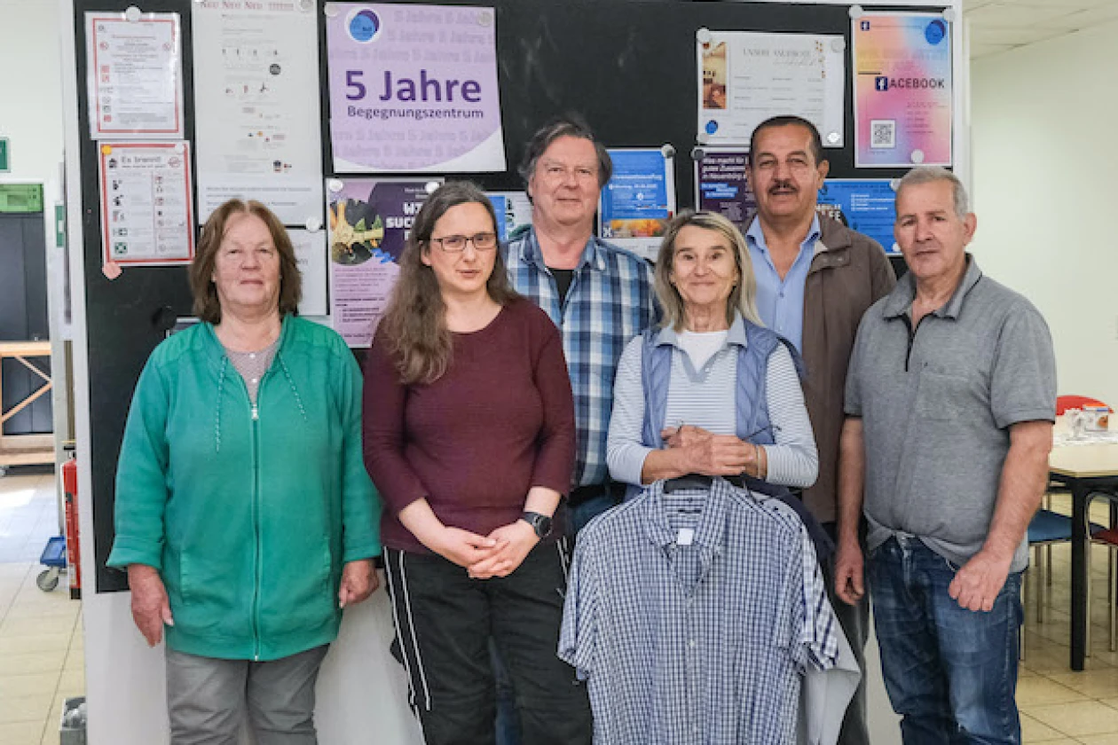 Sechs Personen stehen vor einer Informationswand. Eine Frau h&auml;lt ein kariertes Hemd in der Hand.