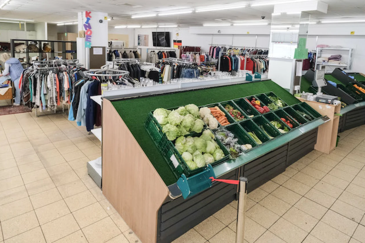 Im Vordergrund steht ein gef&uuml;lltes Gem&uuml;se- und Obstregal, im Hintergrund stehen mit bunten Kleidern best&uuml;ckte Kleiderst&auml;nder und eine arbeitende Frau.