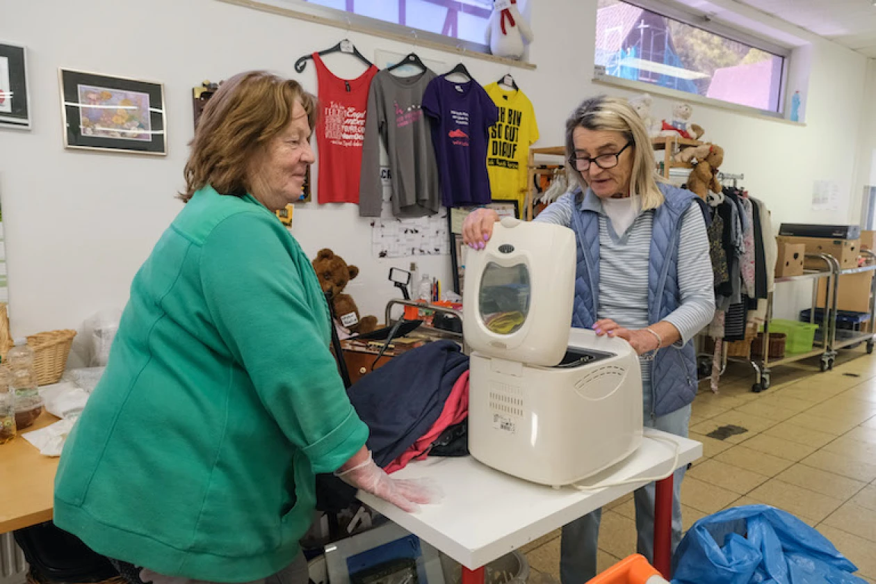 Zwei Frauen in einem Raum am Arbeitstisch. Kleidungsst&uuml;cke, Stofftiere und Haushaltsger&auml;te werden sortiert.