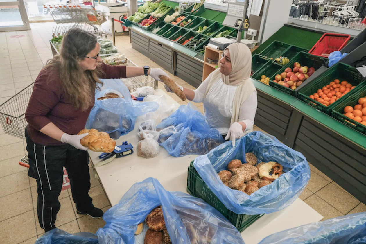 Zu sehen sind zwei Frauen in einem Lebensmittelgesch&auml;ft mit Backwaren in blauen Plastikt&uuml;ten, umgeben von Regalen mit Obst und Gem&uuml;se. Zwischen den zwei Frauen steht ein Tisch, an denen sie die Backwaren mit Handschuhen f&uuml;r den Verkauf vorbereiten.