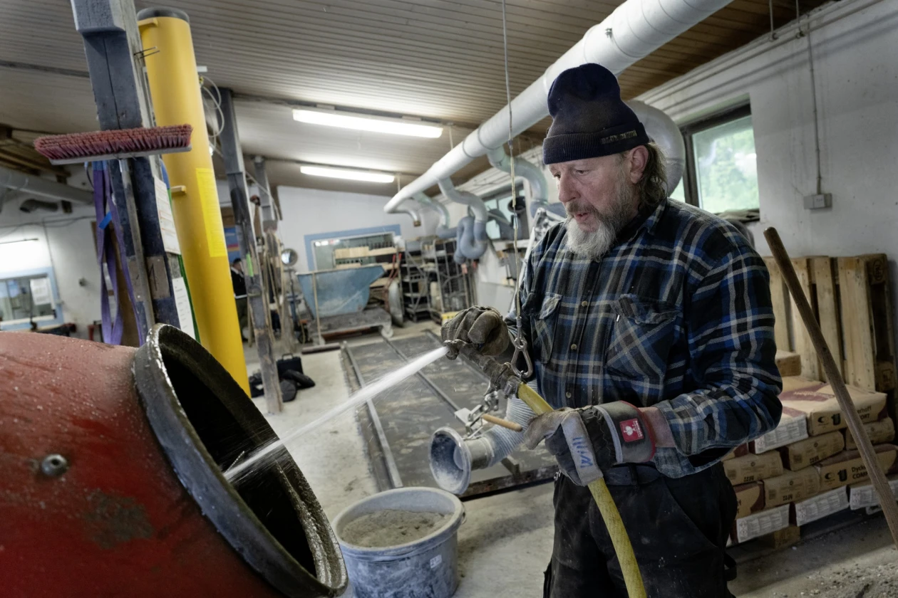 Ein Mann in kariertem Hemd und M&uuml;tze spritz Wasser in einen roten Betonmischer. Im Hintergrund stehen Paletten mit Zements&auml;cken und Werkstattausstattung.