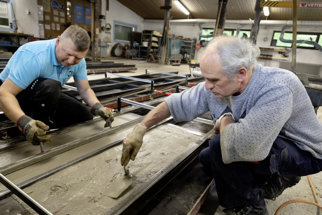 Zwei M&auml;nner bearbeiten in einer Werkstatt ein Betonwerkst&uuml;ck. Der j&uuml;ngere Mann tr&auml;gt ein hellblaues T-Shirt, der &auml;ltere einen grauen Pullover. Beide tragen Arbeitshandschuhe und konzentrieren sich auf ihre Aufgabe.