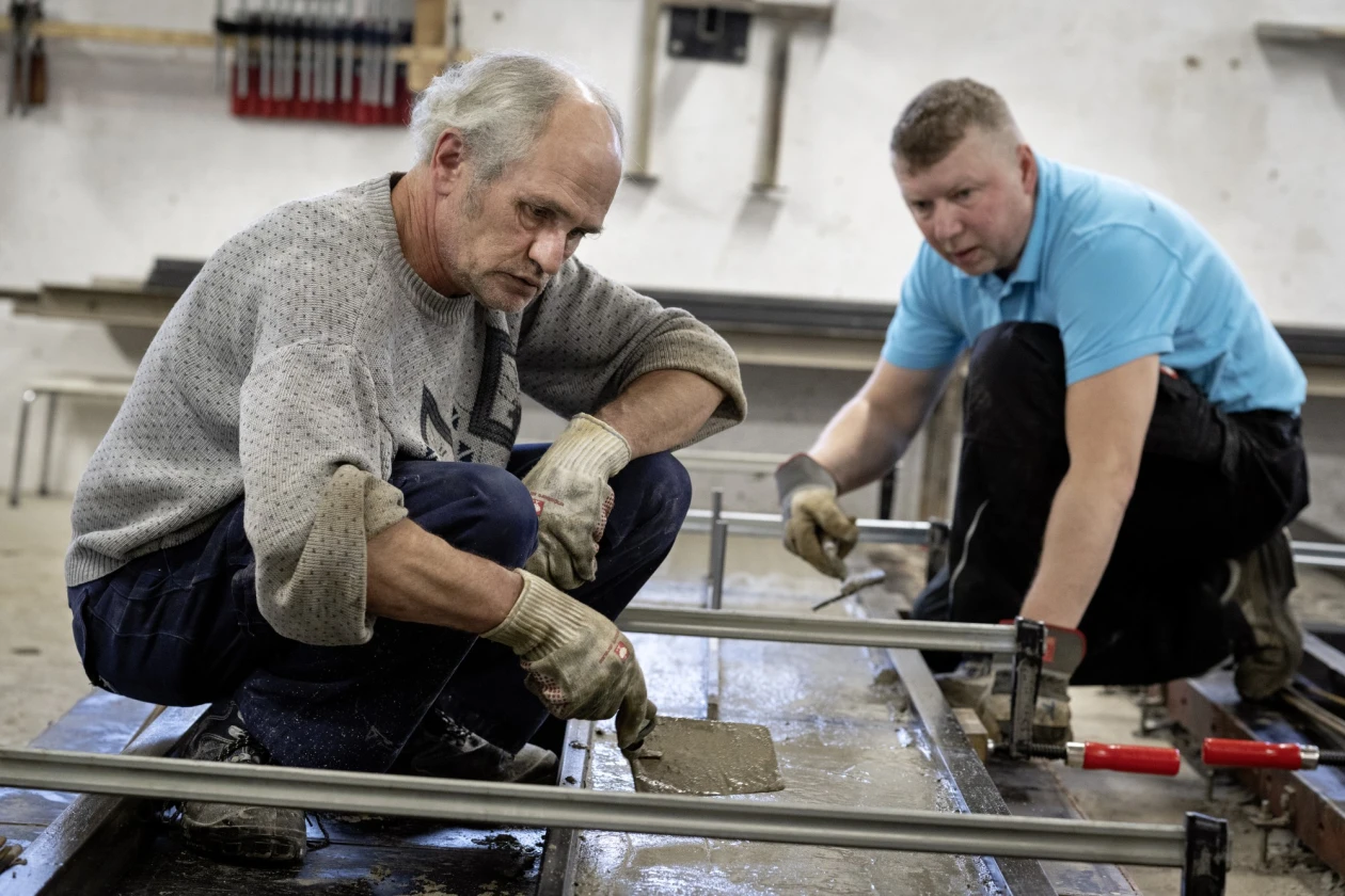 Zwei M&auml;nner bearbeiten ein Betonwerkst&uuml;ck in Schalung. Der j&uuml;ngere Mann tr&auml;gt ein hellblaues T-Shirt, der &auml;ltere einen grauen Pullover. Beide tragen Arbeitshandschuhe und konzentrieren sich auf ihre Arbeit.