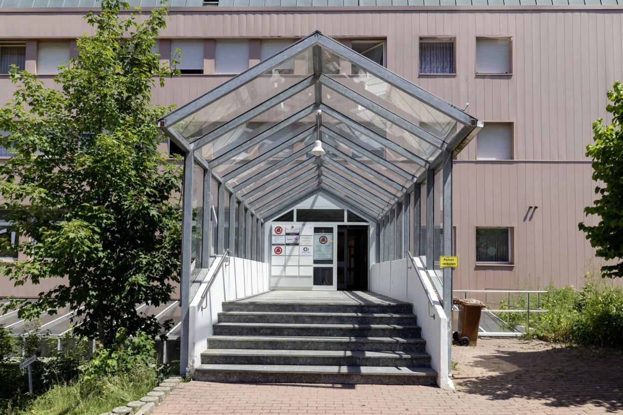 &Uuml;berdachter Eingangsbereich mit Glasdach, Betontreppe und Metallgel&auml;nder, der zu einer Eingangst&uuml;r in einem mehrst&ouml;ckigen Geb&auml;ude f&uuml;hrt.