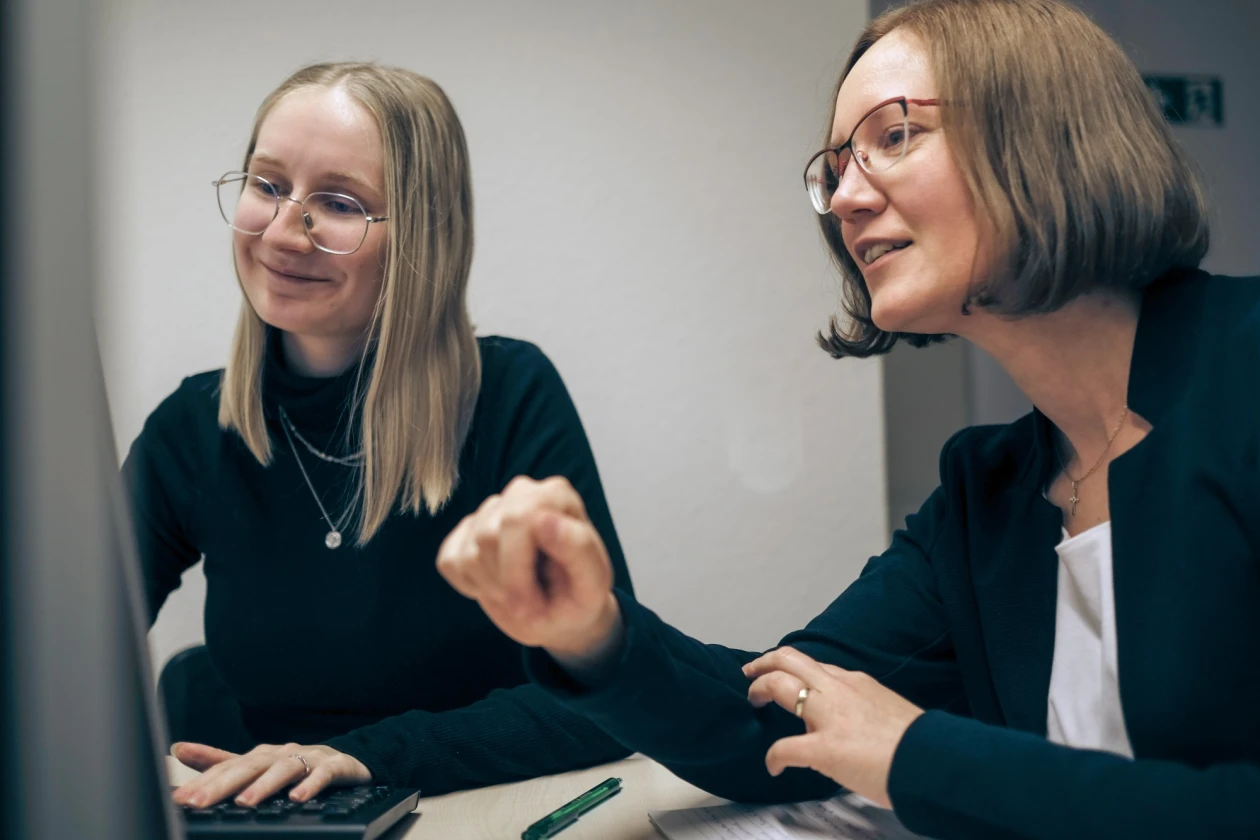 Zwei Frauen sitzen an einem Tisch und schauen in den Computer. Die linke Frau tr&auml;gt schwarze Kleidung, eine Brille und Halsketten. Ihre Hand liegt auf der Tastatur. Die andere Frau tr&auml;gt einen schwarzen Blaser mit einem wei&szlig;en Unterteil und eine Brille.