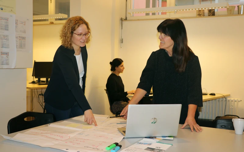 Eine Mitarbeiterin bespricht mit einer Kollegin ausgef&uuml;llte Unterlagen, beide lachen. Im Hintergrund sitzt eine Frau am Computer.