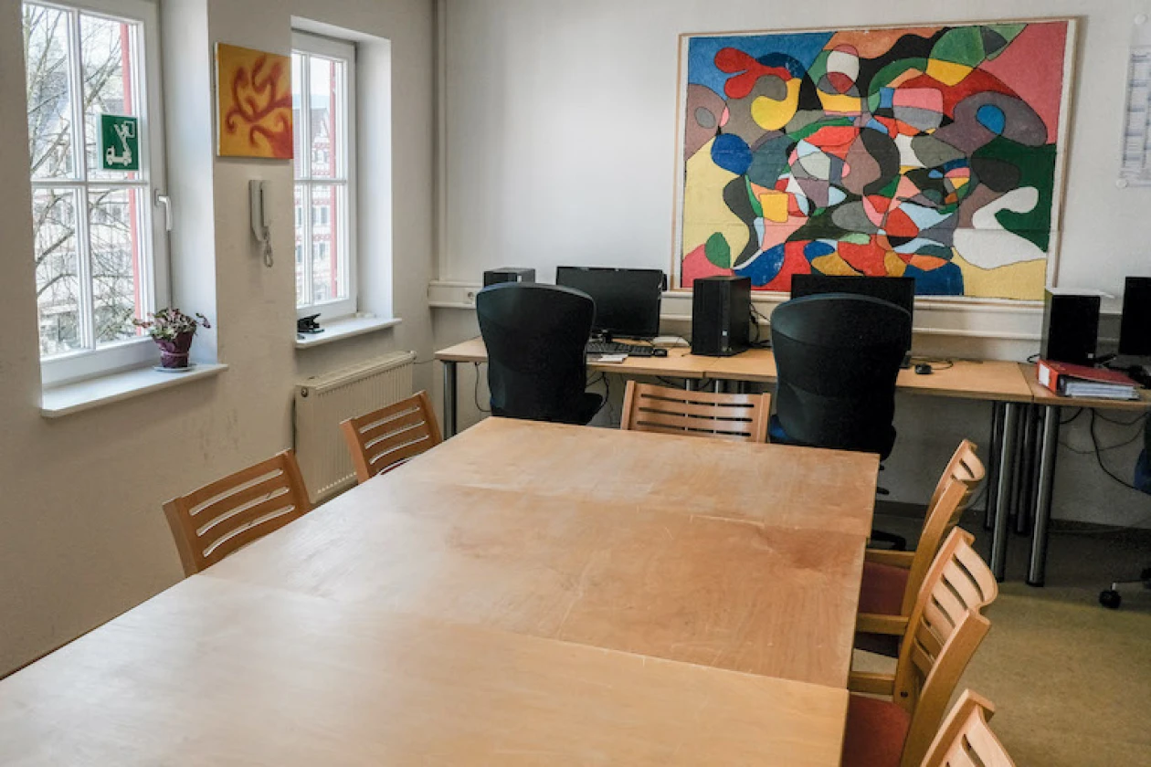 Ein Raum mit zwei Fenstern, einem gro&szlig;em Holztisch, Holzst&uuml;hlen und im Hintergrund PC-Arbeitspl&auml;tze mit schwarzen Schreibtischst&uuml;hlen. Ein buntes, abstraktes Kunstwerk befindet sich an der Wand. Eine kleine Pflanze steht auf der Fensterbank.