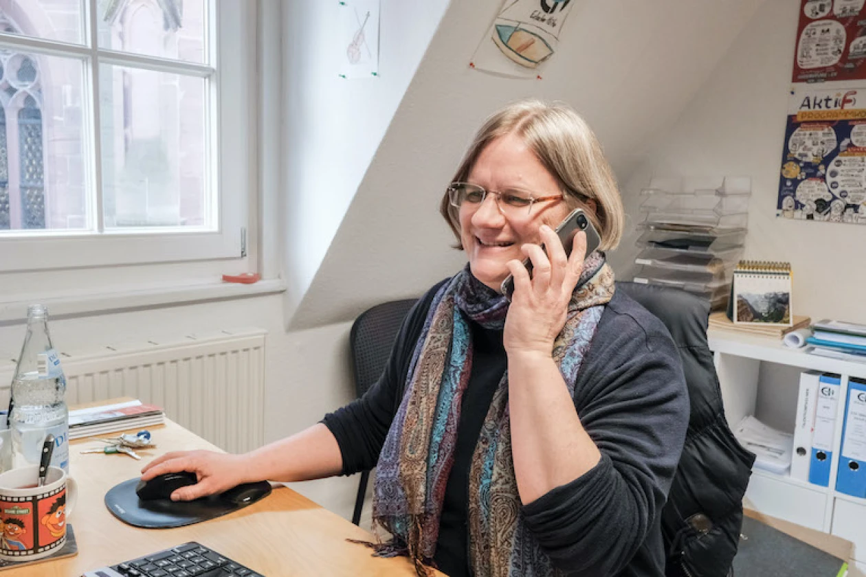Eine Frau mit Brille sitzt an ihrem Schreibtisch in einem hellen Dachzimmer und telefoniert