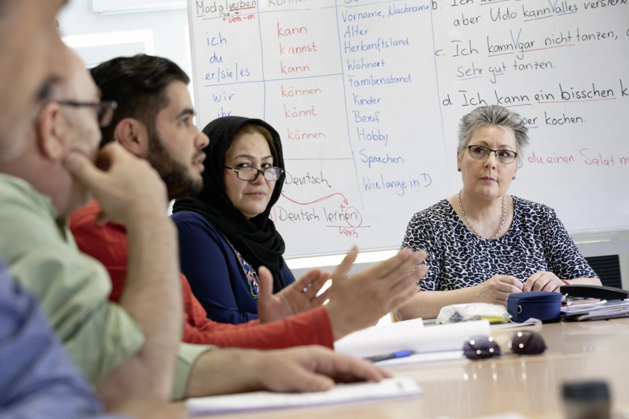Eine Frau in gemustertem T-Shirt und Brille sitzt frontal am Tisch. Neben ihr sind weitere Personen zu erkennen, die gestikulieren und aufmerksam zuh&ouml;ren. Im Hintergrund ist eine Tafel mit Sprachlernmaterialien zu erkennen. Die Atmosph&auml;re wirkt konzentriert und lernorientiert.