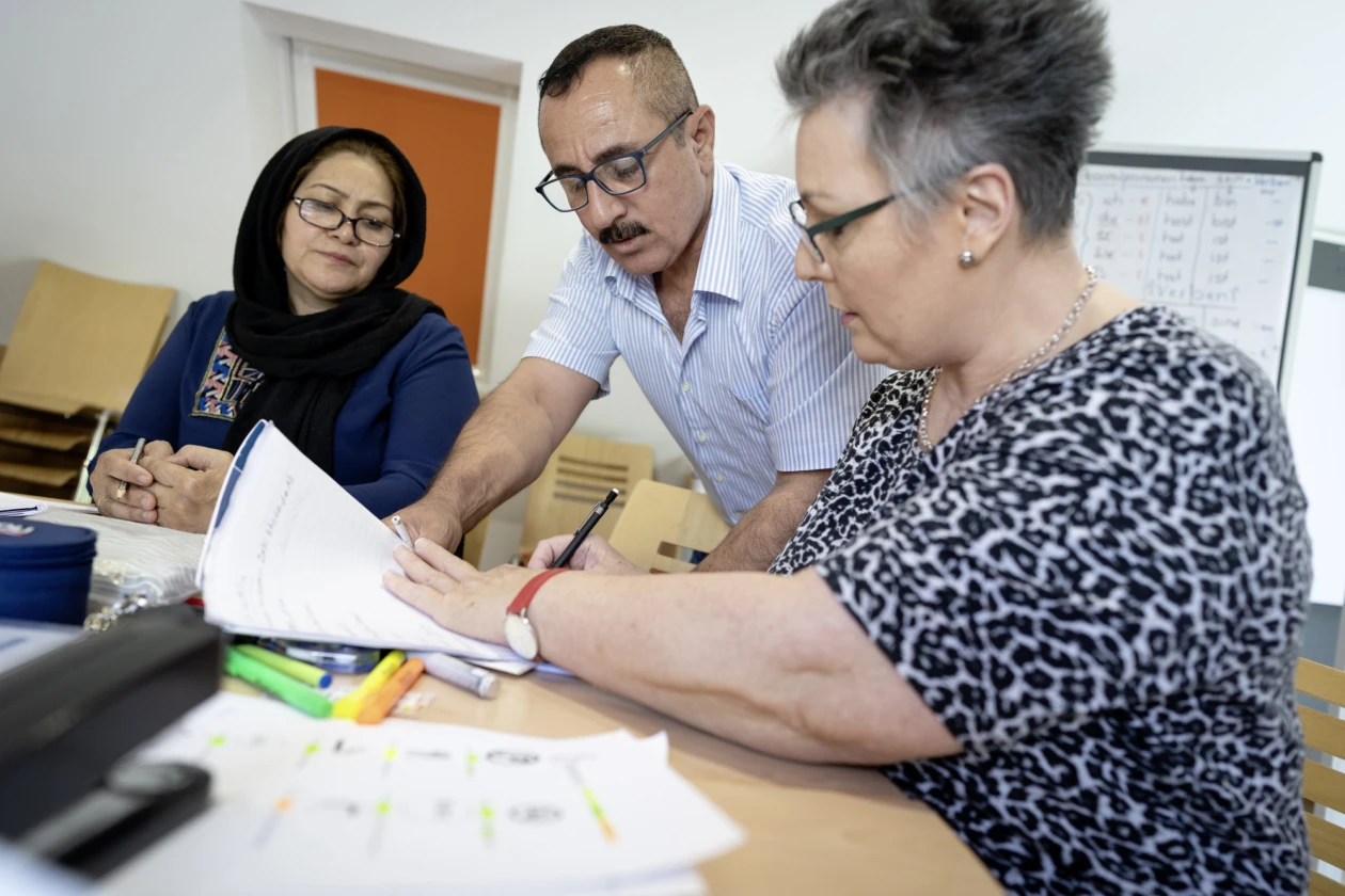 Drei Personen sitzen an einem Tisch. Die Frau in gemustertem T-Shirt und Brille h&auml;lt ein Dokument in der Hand und erkl&auml;rt etwas, w&auml;hrend der Mann mit kariertem Hemd und Brille, sowie die Frau mit Kopftuch und ebenfalls einer Brille auf das Dokument schauen und zuh&ouml;ren.