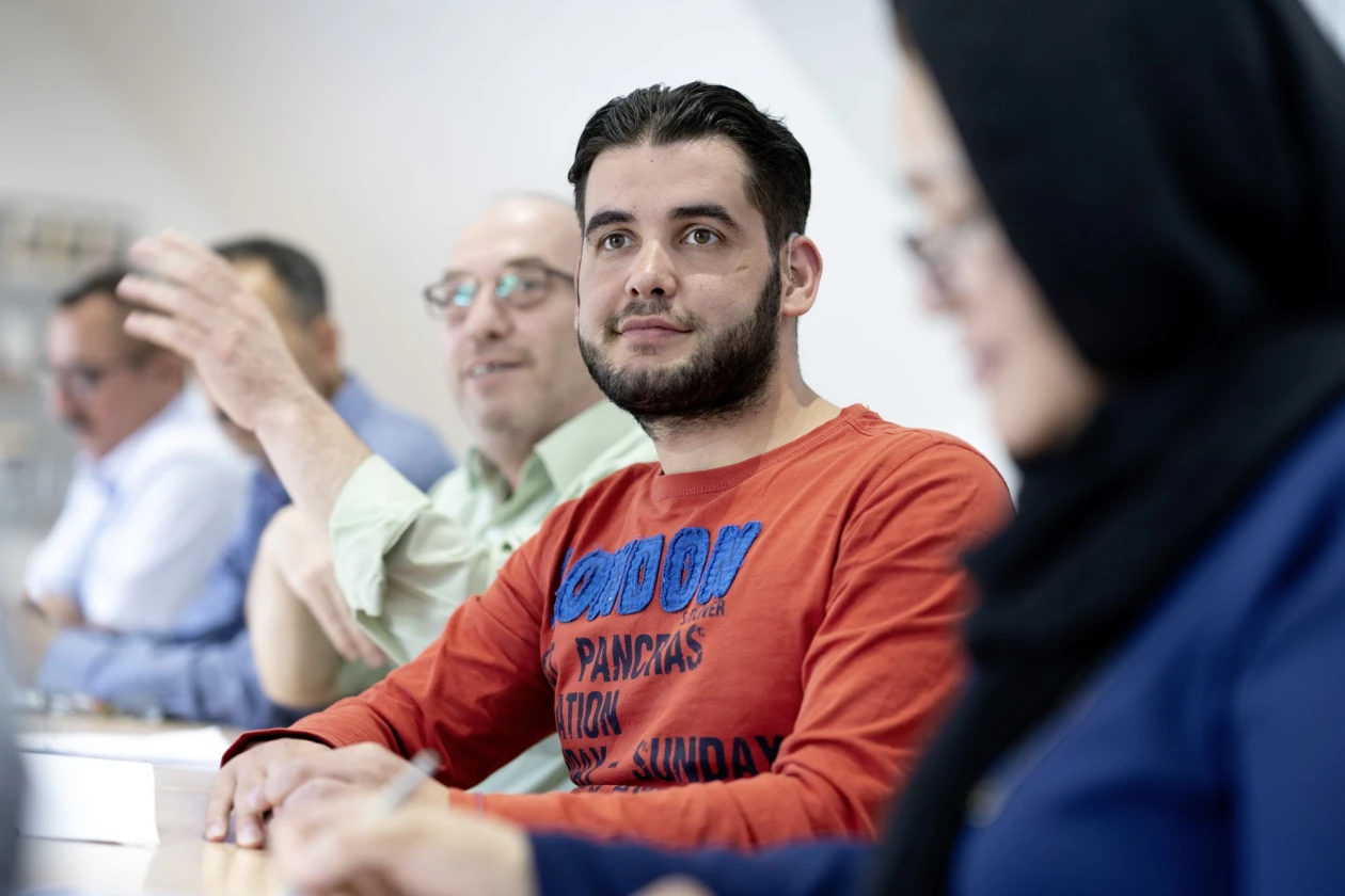 Ein junger Mann im orangefarbenen Pullover sitzt in einem Seminarraum und blickt konzentriert nach vorne, w&auml;hrend im Hintergrund weitere Teilnehmer sichtbar sind.