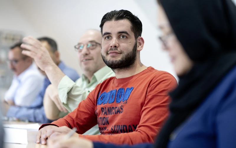 Ein junger Mann im orangefarbenen Pullover sitzt in einem Seminarraum und blickt konzentriert nach vorne, w&auml;hrend im Hintergrund weitere Teilnehmer sichtbar sind.