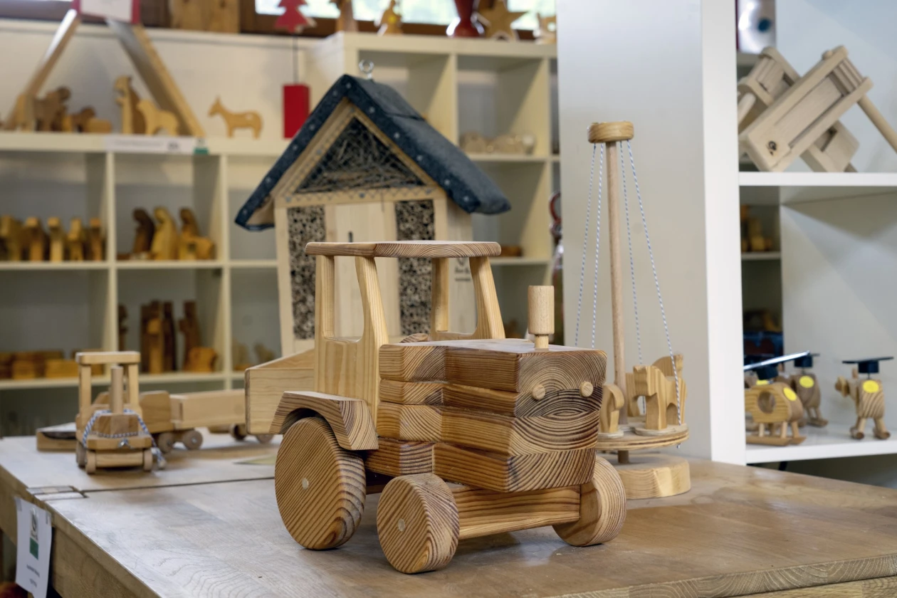 Handgefertigter Spielzeug-Traktor aus Holz mit Details wie R&auml;dern und Kabine auf einem Tisch. Im Hintergrund sieht man weitere handgefertigte Holzspielzeuge.