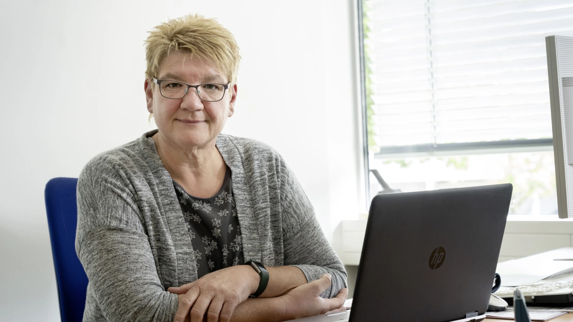 Eine Frau mit kurzen blonden Haaren und Brille sitzt an einem Schreibtisch mit einem Laptop. Sie tr&auml;gt einen grauen Cardigan &uuml;ber einem gebl&uuml;mten Oberteil und l&auml;chelt freundlich in die Kamera. Der Arbeitsplatz befindet sich in einem hellen B&uuml;roraum mit Fenster im Hintergrund.