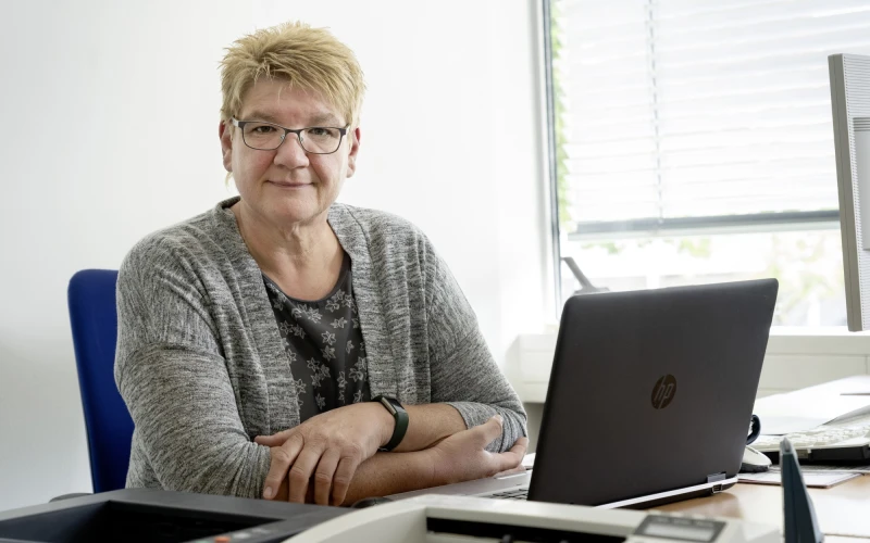 Eine Frau mit kurzen blonden Haaren und Brille sitzt an einem Schreibtisch mit einem Laptop. Sie tr&auml;gt einen grauen Cardigan &uuml;ber einem gebl&uuml;mten Oberteil und l&auml;chelt freundlich in die Kamera. Der Arbeitsplatz befindet sich in einem hellen B&uuml;roraum mit Fenster im Hintergrund.