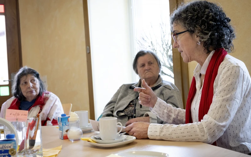 Drei Frauen sitzen an einem Tisch mit Kaffeegedecken. Die Frau im wei&szlig;en Pullover und rotem Schal erz&auml;hlt, w&auml;hrend die anderen beiden aufmerksam zuh&ouml;ren.