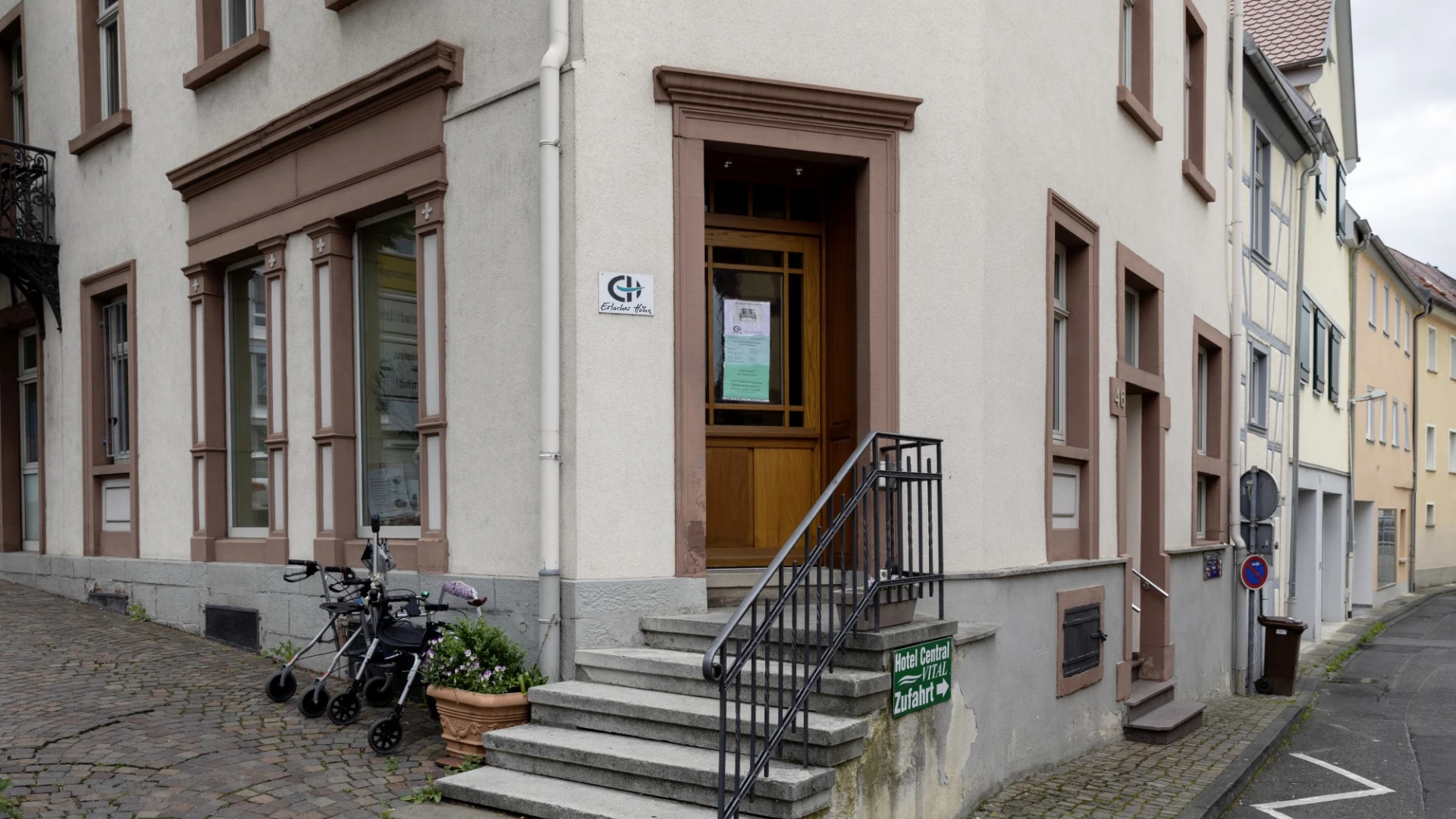 Eingangstreppe eines &auml;lteren Geb&auml;udes mit Gel&auml;nder und einem Schild mit dem Logo der Erlacher H&ouml;he. Neben der Eingangstreppe stehen zwei geparkte Rollatoren.