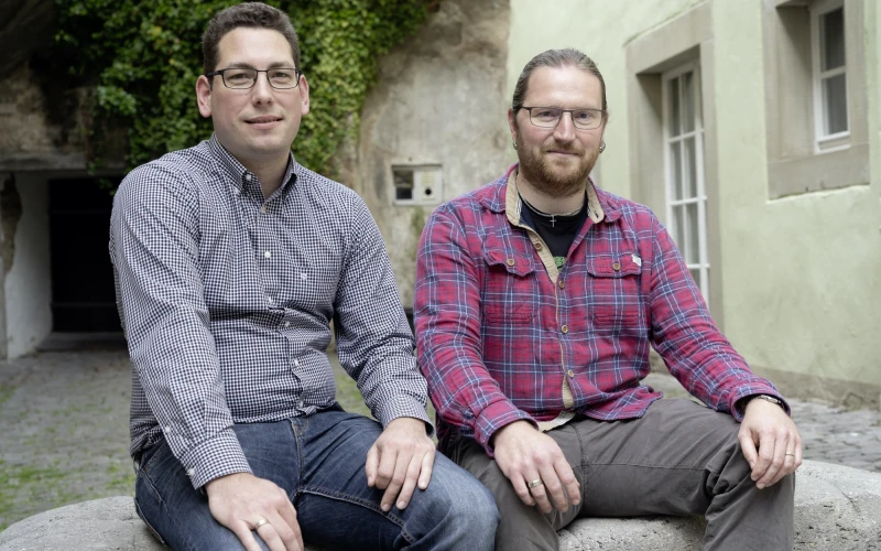 Zwei M&auml;nner sitzen nebeneinander vor einem Geb&auml;ude. Der linke tr&auml;gt ein kleinkariertes Hemd und eine Brille, der rechte tr&auml;gt ein rotes Karohemd und tr&auml;gt ebenfalls eine Brille. Sie blicken l&auml;chelnd in die Kamera.