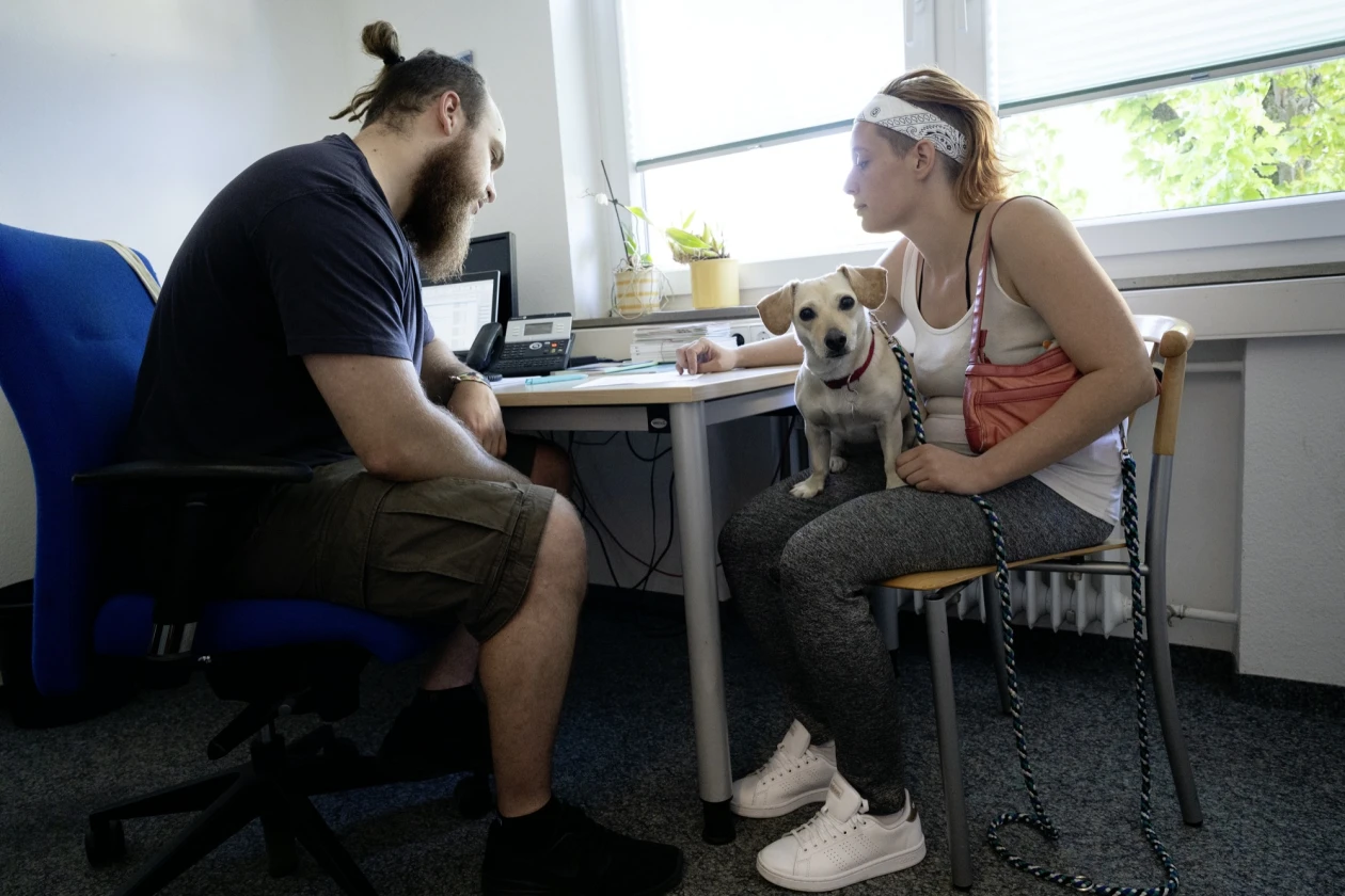 Ein Mann und eine Frau sitzen an einem Schreibtisch. Zwischen ihnen sitzt ein Hund. Sie befinden sich in einem hellen B&uuml;roraum mit Fenster, einem Computer und weiteren B&uuml;romaterialien.