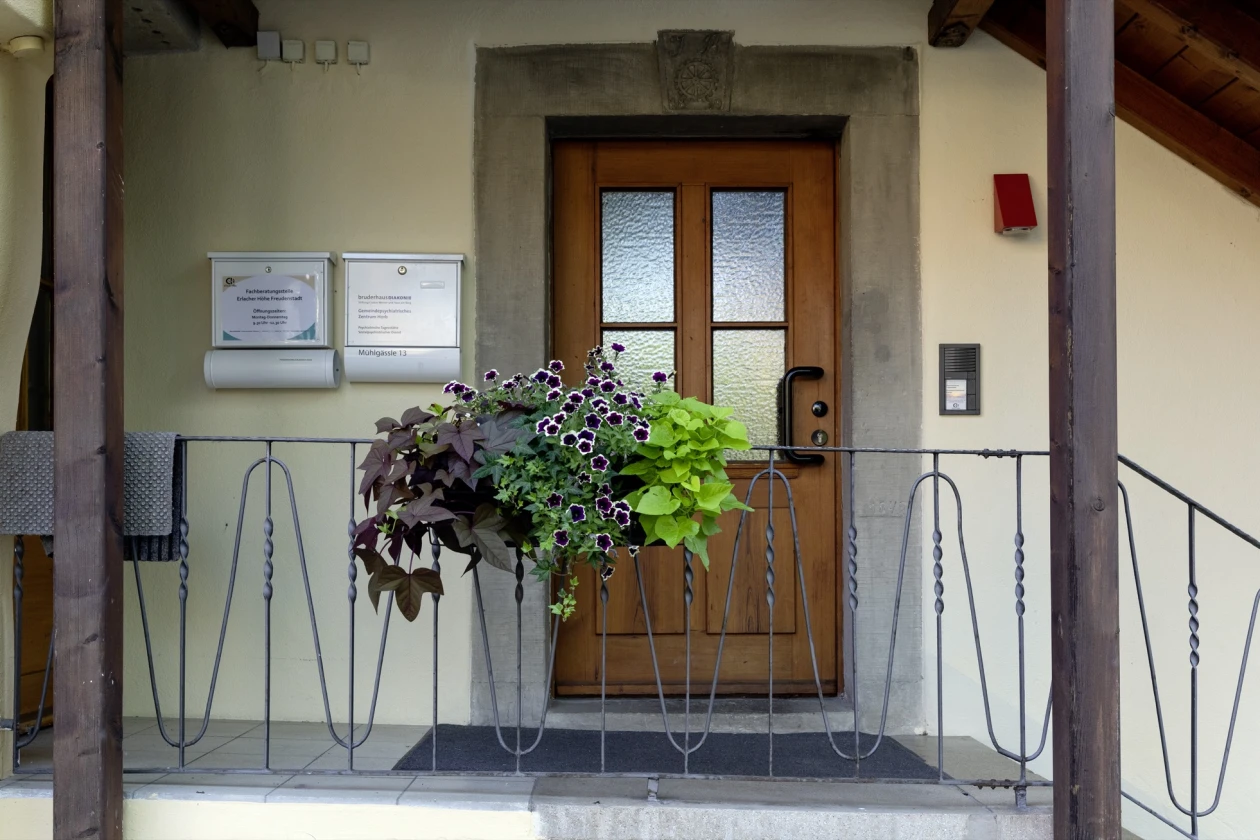 Eine Holzt&uuml;r mit Milchglaselementen im Eingangsbereich, flankiert von einem Metallgel&auml;nder und einem Blumenarrangement am Gel&auml;nder h&auml;ngend. Zwei Briefk&auml;sten h&auml;ngen links neben der T&uuml;r an der Wand.