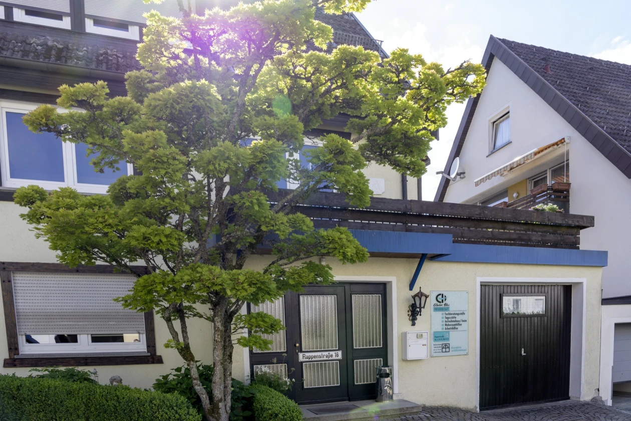 Ein wei&szlig;es Geb&auml;ude mit Garage. Im Vordergrund ein Baum mit gr&uuml;nen Bl&auml;ttern und eine kleine Hecke. Rechts von der Haust&uuml;re h&auml;ngt eine Informationstafel der Erlacher H&ouml;he.