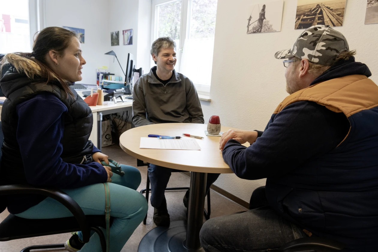 Drei Personen sitzen an einem runden Tisch in einem B&uuml;ro mit Fenster. Eine Frau und zwei M&auml;nner unterhalten sich miteinander, w&auml;hrend Papiere und Kulis auf dem Tisch liegen. Der Raum ist hell und mit Bildern an den W&auml;nden dekoriert.