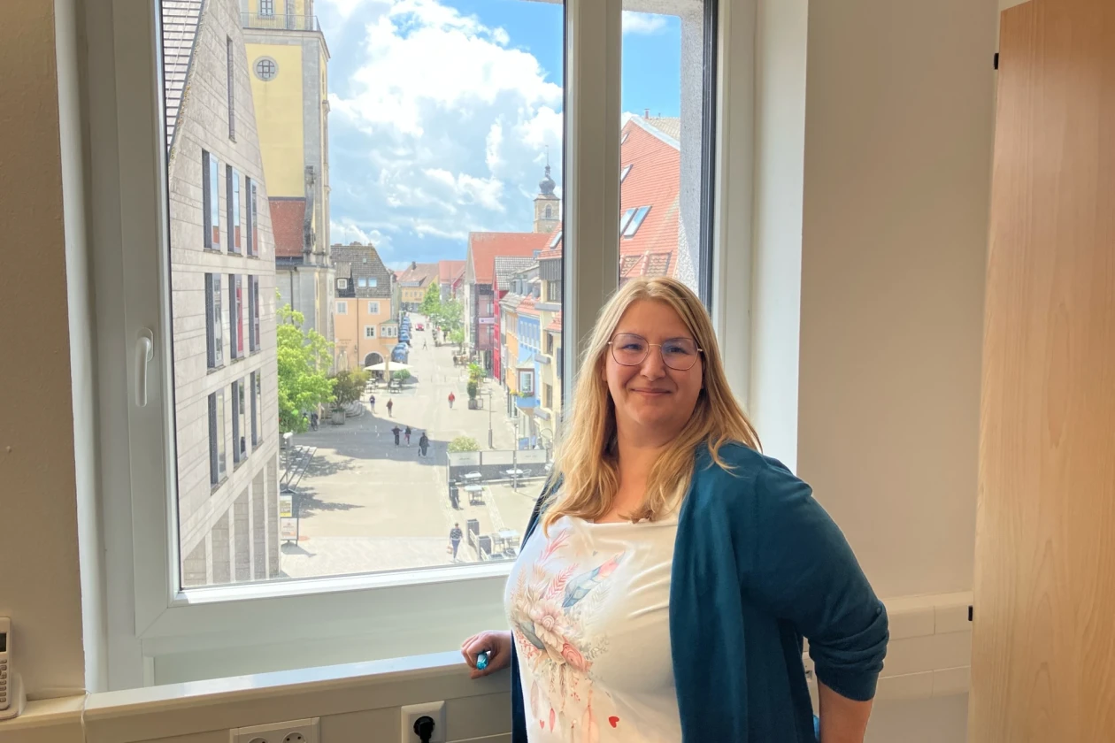 Frau mit langen blonden Haaren, Brille und blauem Cardigan steht vor einem ge&ouml;ffneten Fenster mit Blick auf eine belebte Altstadtstra&szlig;e.