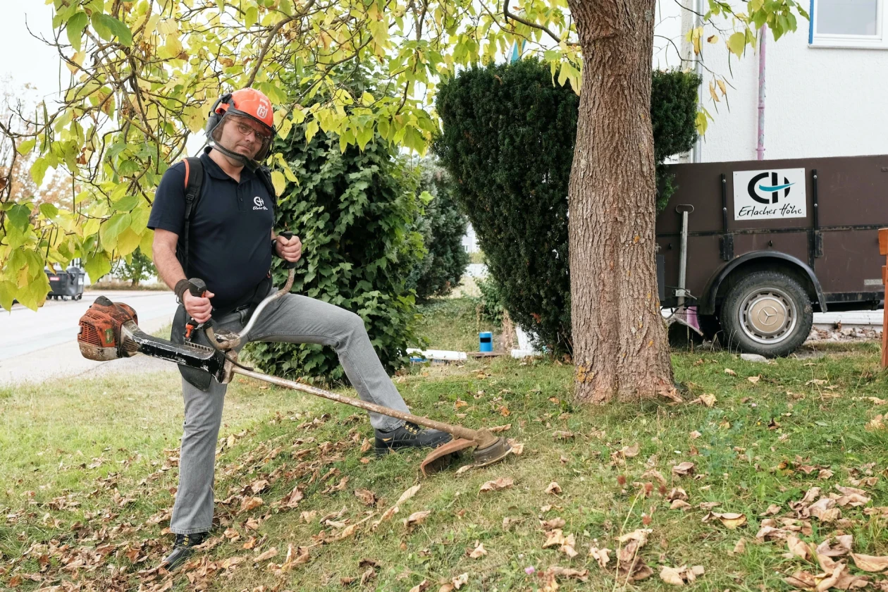 Mann mit Motorsense steht auf einem grasbewachsenen Hang neben einem Baum und schneidet Gras. Im Hintergrund ist ein Anh&auml;nger mit dem Logo der Erlacher H&ouml;he zu sehen.